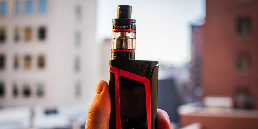 a hand holding a black and red vape device with buildings in the background